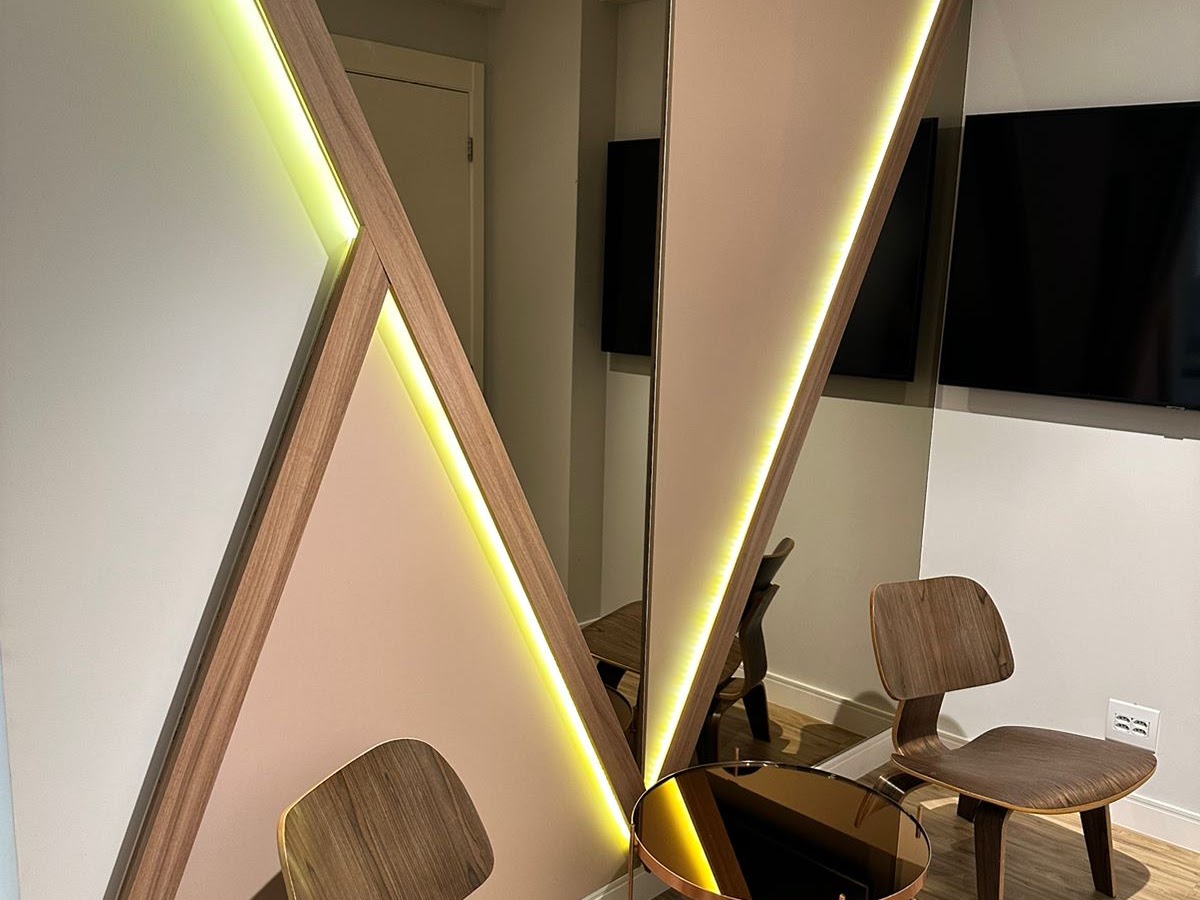 Recepção do consultório com painel geométrico de madeira iluminado por fitas de LED, espelho e cadeiras de madeira estofadas